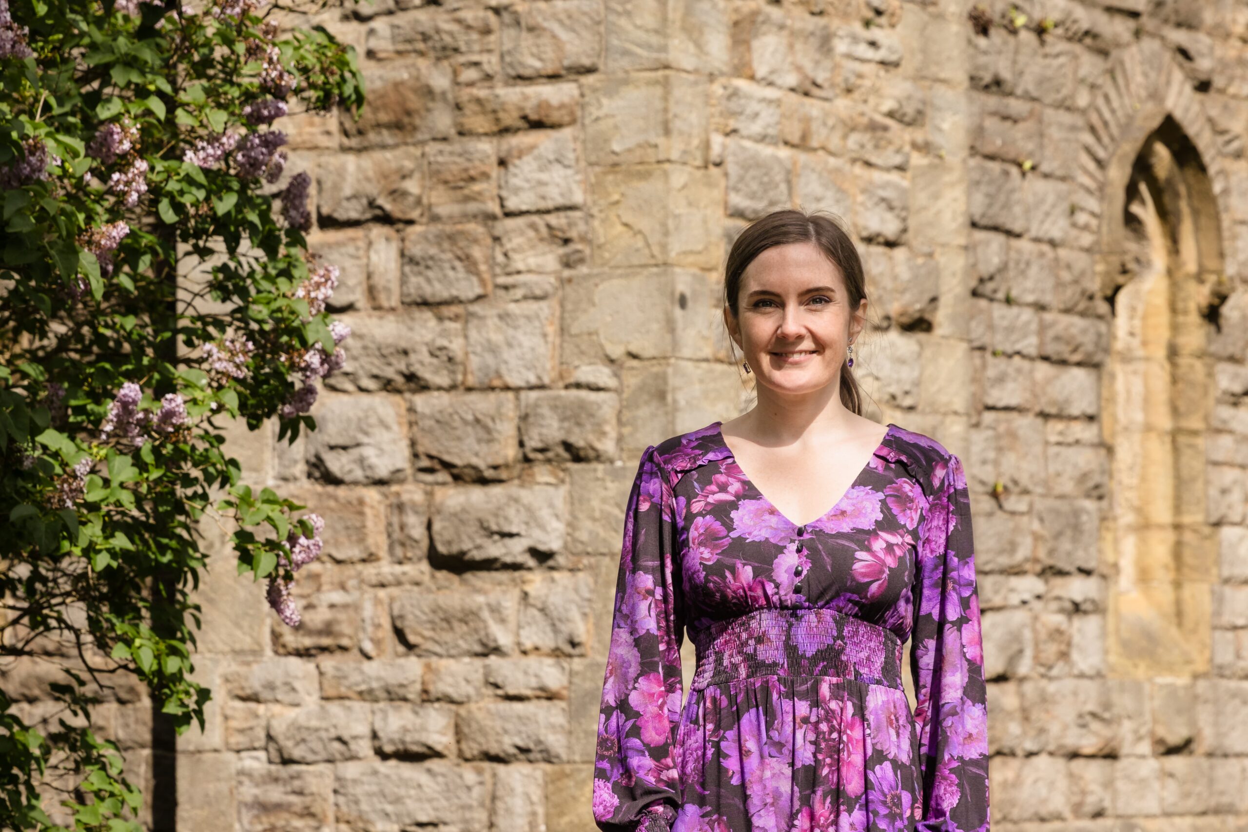 Emma McClelland, an independent celebrant, stands in front of a church in a purple floral dress