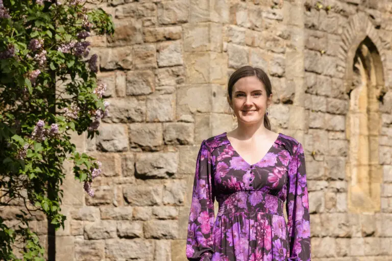 Emma, an independent celebrant, is standing in front of a church wearing a purple floral dress