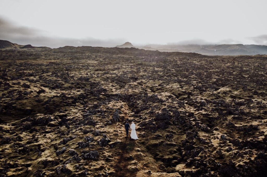 Couple in wedding attire walking across a vast, rocky Icelandic landscape at sunset.